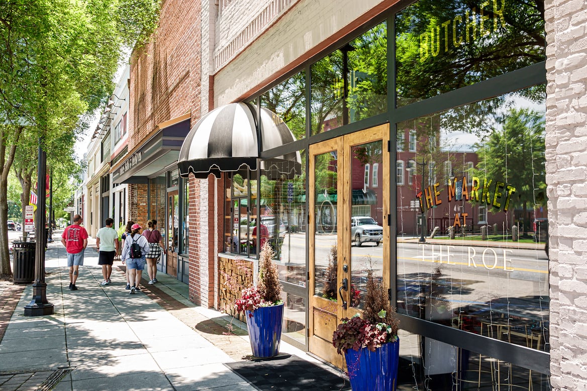 A group of people walk down a street with multiple storefronts on a sunny day.